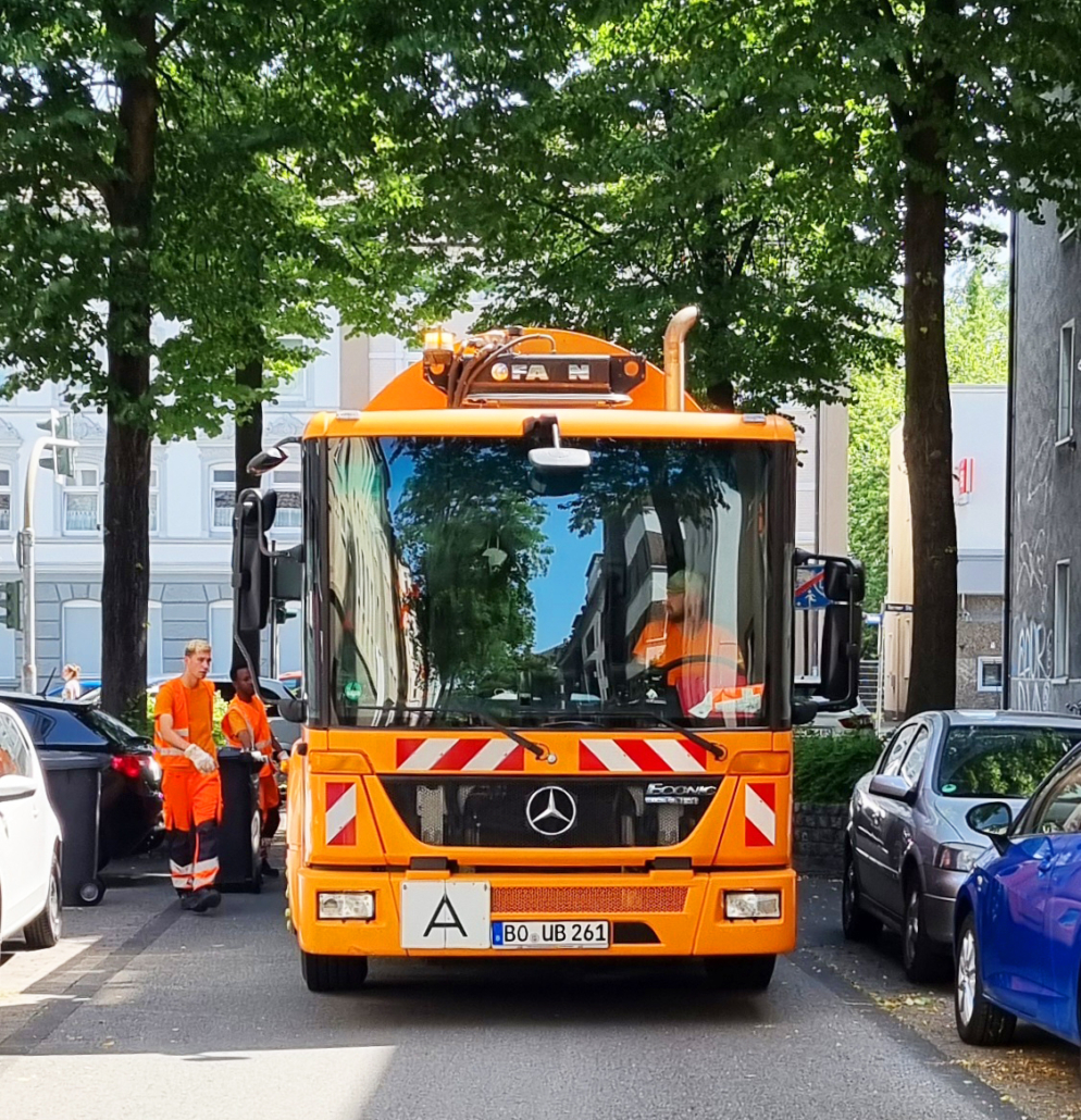 Ein Müllauto von vorne in einer schmalen Straße, am Rand sind Bäume