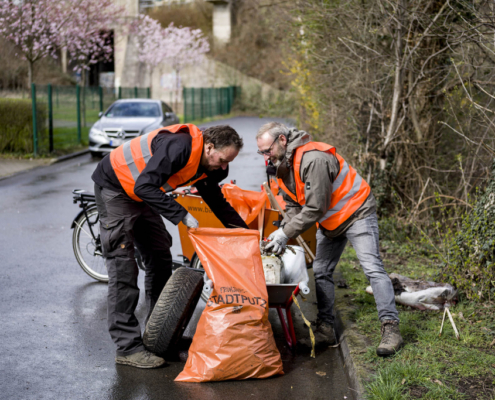 Zwei Männer mit Warnweste packen Müll am Straßenrand in einen Stadtputzsack.