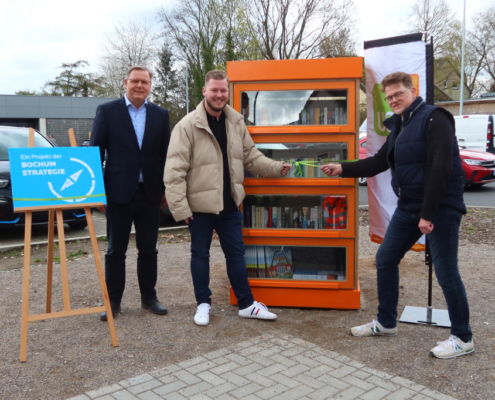 Vor einem Bücherschrank stehen 3 Männer, 2 von Ihnen zeihen eine Grüne Schleife auf, welche um den Bücherschrank gebunden ist. an der linken Seite ist ein blaues Schild mit der Aufschrift Ein Projekt der Bochum Strategie zu sehen.