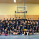 Mehrere Kinder und Betreuer stehen in einer Sporthalle unter einem Basketballkorb.