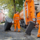 2 Tonnen werden von Mitarbeitern zum Müllwagen gezogen. der Müllwagen steht am Straßenrand und ist von hinten abgebildet.