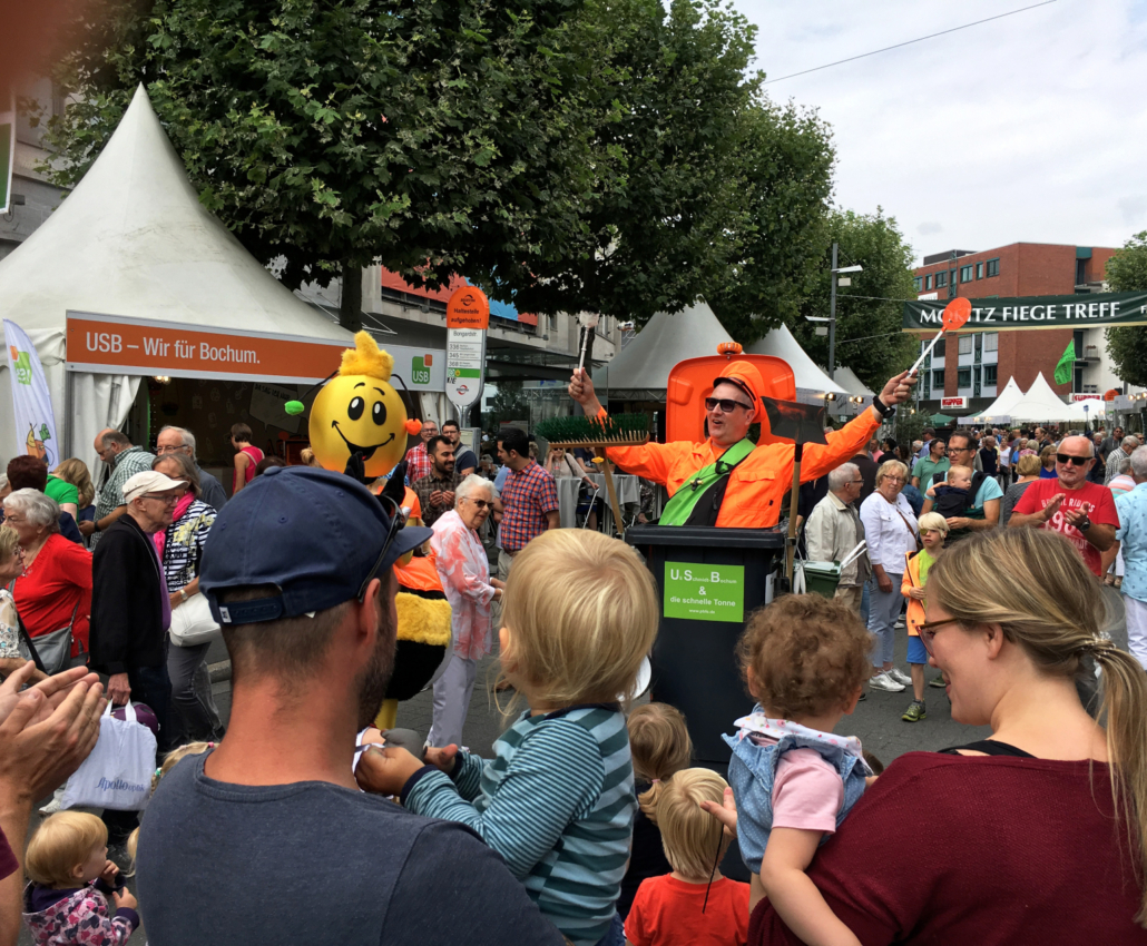 Ein Mann gekleidet als Mülleimer wird von Außenstehenden applaudiert