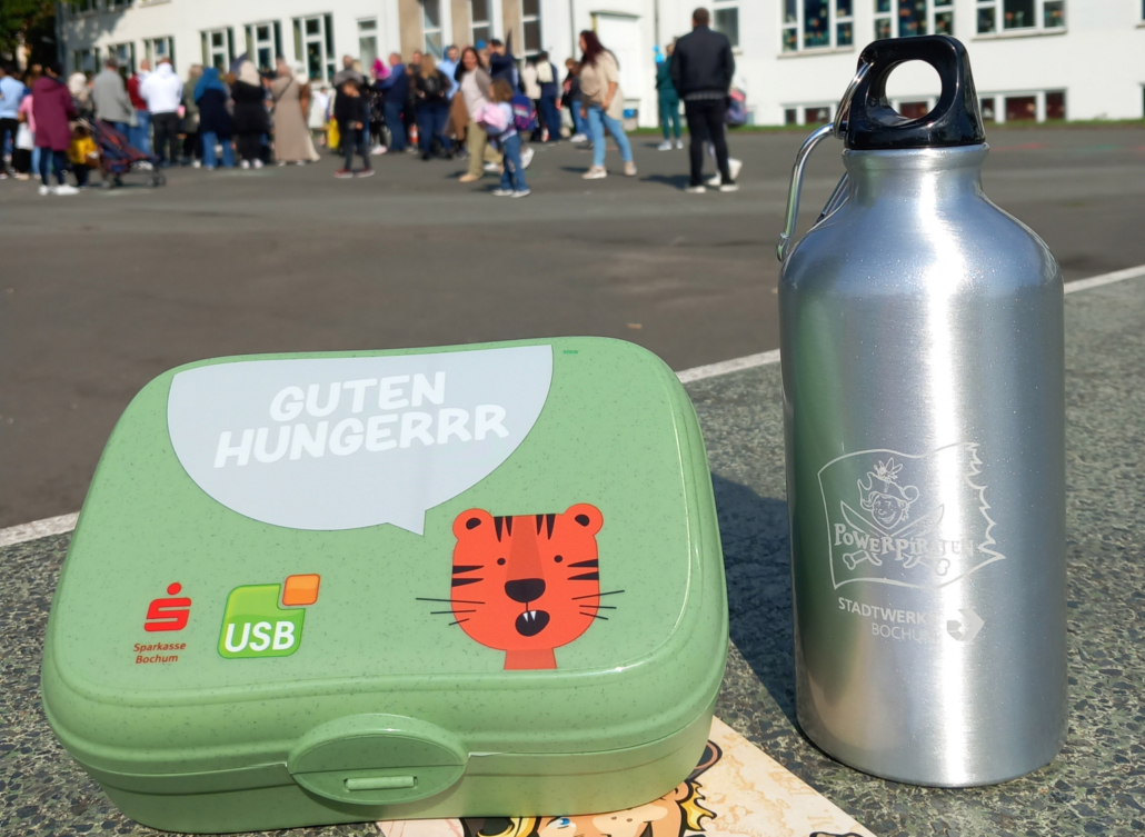 Eine Brotdose des USBs und eine Flasche der Powerpiraten stehen auf einem Schulhof. Im Hintergrund sind Schüler zu erkennen