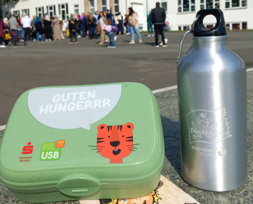 Eine Brotdose des USBs und eine Flasche der Powerpiraten stehen auf einem Schulhof. Im Hintergrund sind Schüler zu erkennen
