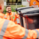 Ein lächelnder Mitarbeiter lädt zusammen mit seinem Kollegen Mülleimer in den Müllwagen ein