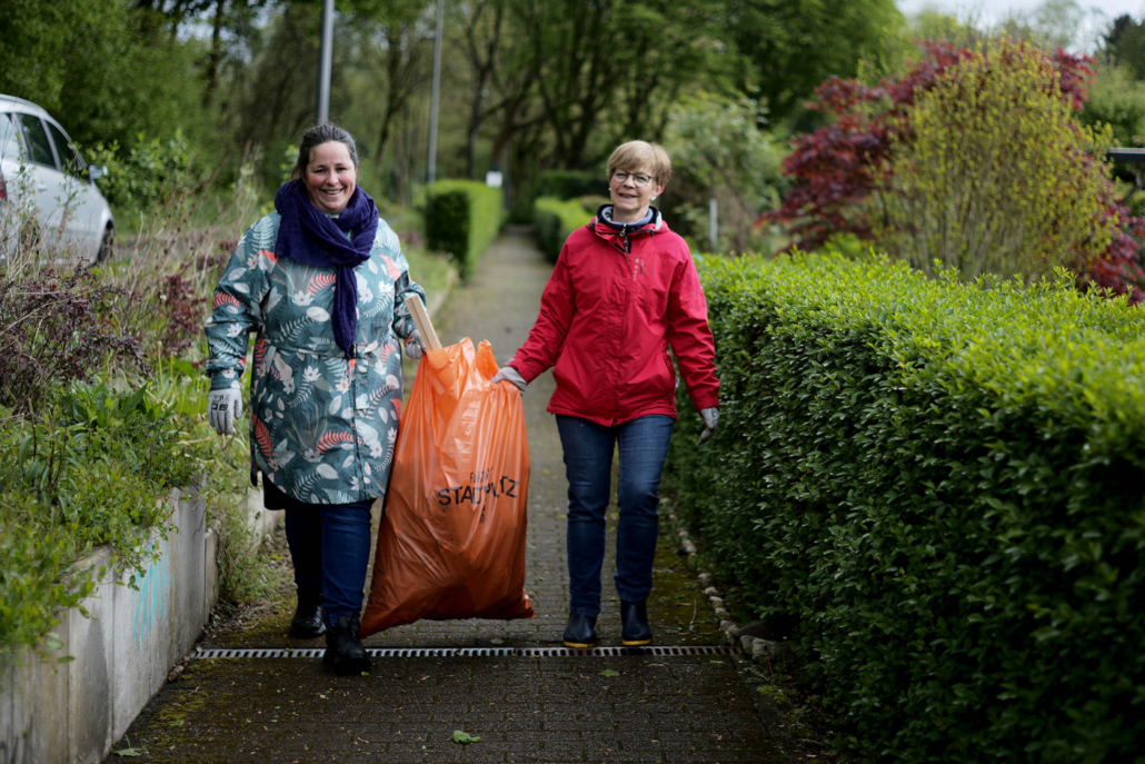 Zwei Frauen tragen einen orangenen Müllsack. sie befinden Sich auf einem Gehweg umrandet mit Büschen.
