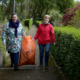 Zwei Frauen tragen einen orangenen Müllsack. sie befinden Sich auf einem Gehweg umrandet mit Büschen.