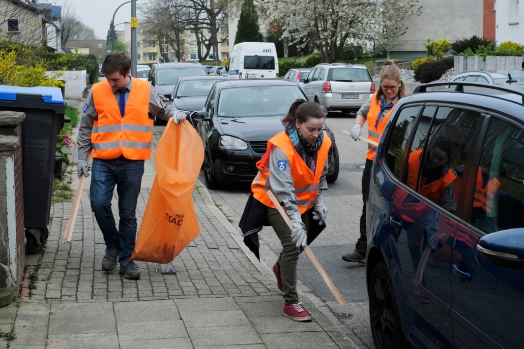 Drei Menschen heben Müll auf der Bürgersteig mit Zangen auf und tragen orangene warnwesten. einer davon hält einen orangenen Stadtputzsack in der Hand. am Straßenrand parken Autos