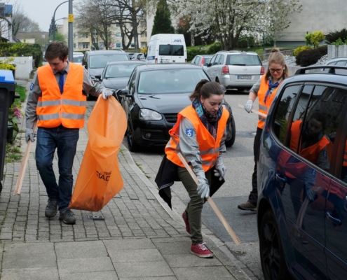 Drei Menschen heben Müll auf der Bürgersteig mit Zangen auf und tragen orangene warnwesten. einer davon hält einen orangenen Stadtputzsack in der Hand. am Straßenrand parken Autos