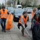 Drei Menschen heben Müll auf der Bürgersteig mit Zangen auf und tragen orangene warnwesten. einer davon hält einen orangenen Stadtputzsack in der Hand. am Straßenrand parken Autos