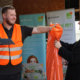 ein junger Mann mit einem Blauen Klemmbrett in der Hand übergibt einen orangenen Stadputzsack an eine junge Blonde Frau