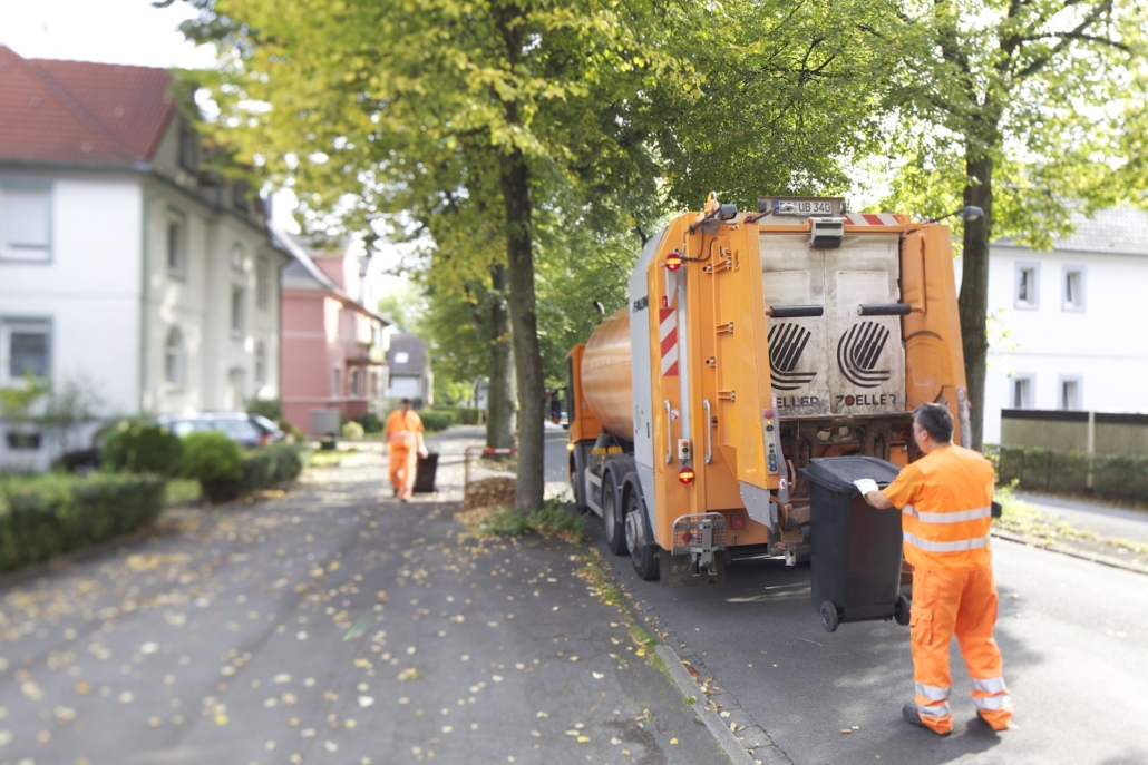 Ein Mitarbeiter in orangener Arbeitskleidung hängt eine Tonne an den Müllwagen, im Hintergrund zieh ein anderer eine Tonne zum Müllwagen.