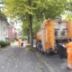 Ein Mitarbeiter in orangener Arbeitskleidung hängt eine Tonne an den Müllwagen, im Hintergrund zieh ein anderer eine Tonne zum Müllwagen.