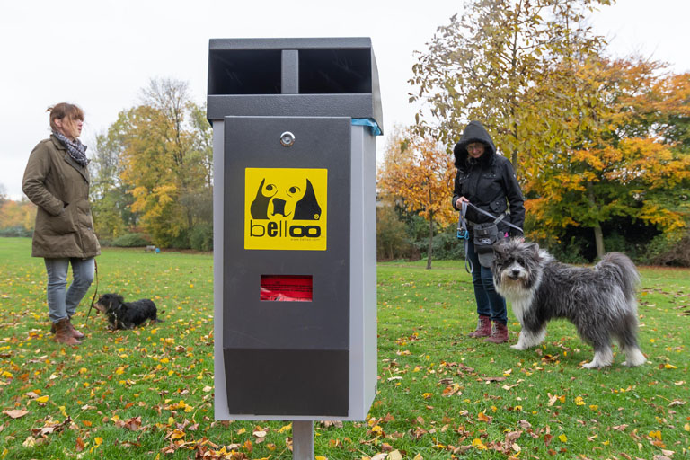 Hundekotbeutelspender im Vordergrund. Daneben stehen Hunde mit ihren Besitzern