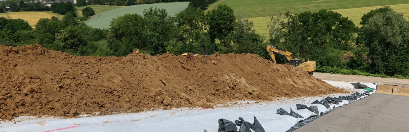 Erdhaufen liegen auf der Deponie. Im Hintergrund sieht man einen Bagger