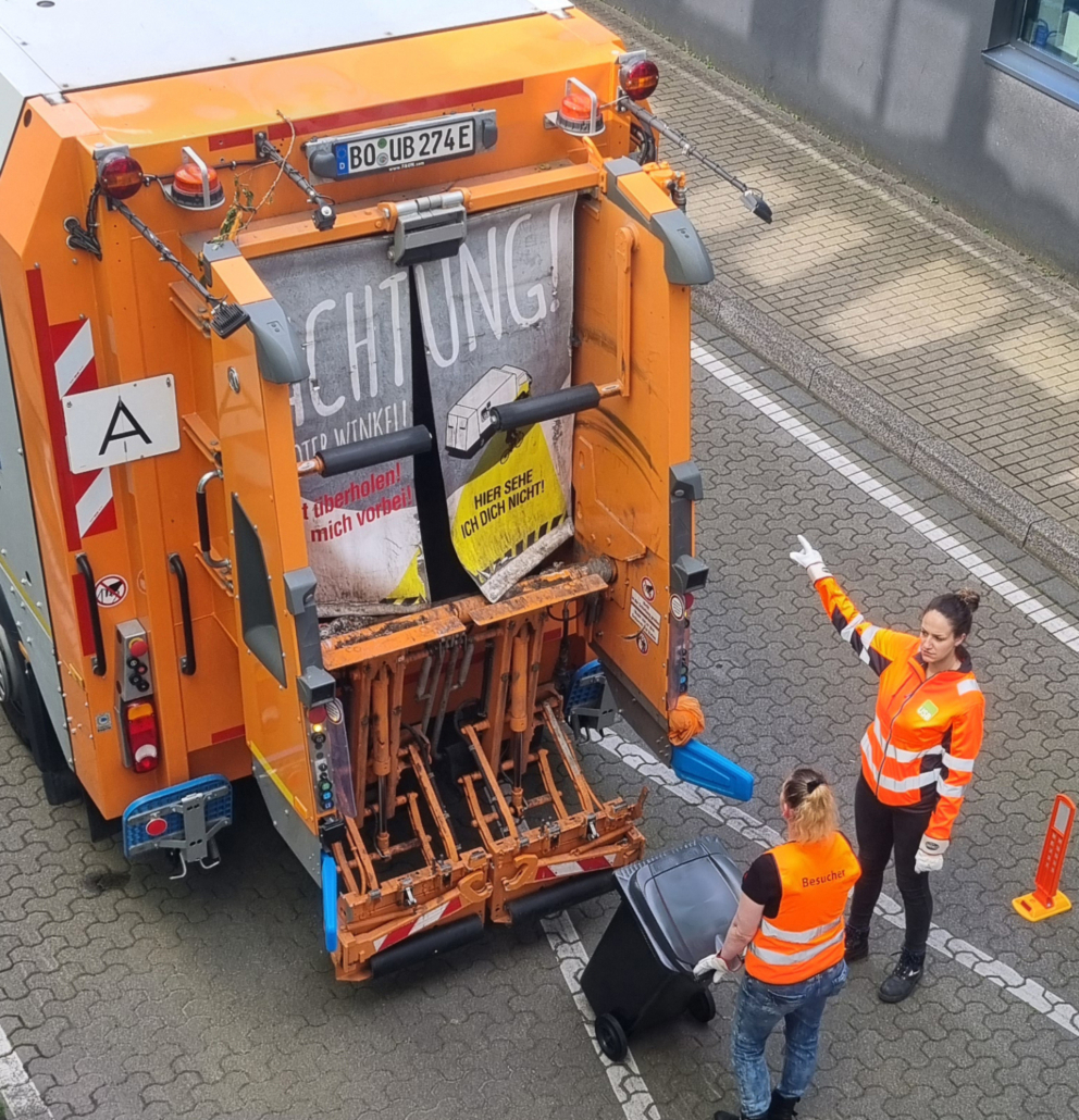 Eine junge Frau erklärt einer anderen, wie eine Tonne am Müllauto einzuhängen ist. vor ihnen steht ein Müllwagen