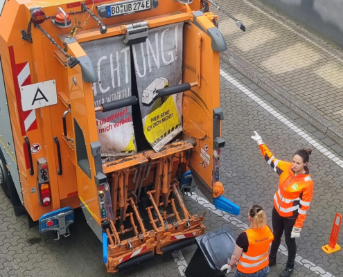 Zwei Frauen in orangenen Westen laden Müll in einen Müllwagen