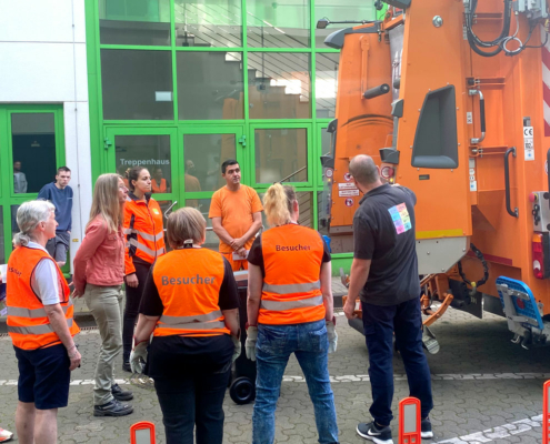 Orange gekleidete Personen stehen hinter einem Müllwagen und beobachten diesen