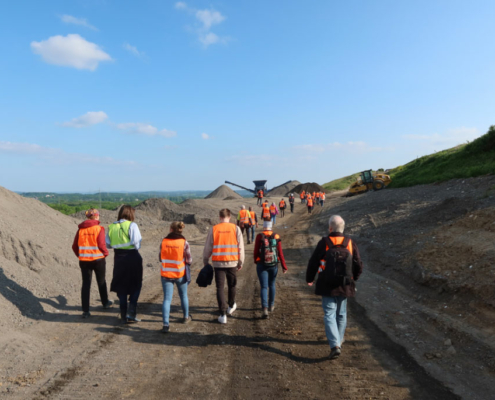 Eine Gruppe von Menschen schaut sich eine Baustelle an. Im Hintergrund ist eine grüne Landschaft und ein Bagger zu sehen