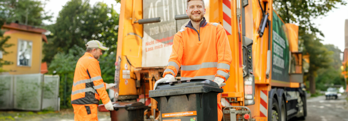 Zwei Männer holen den Müll ab. Sie stehen vor einem Müllwagen und sind orange gekleidet.