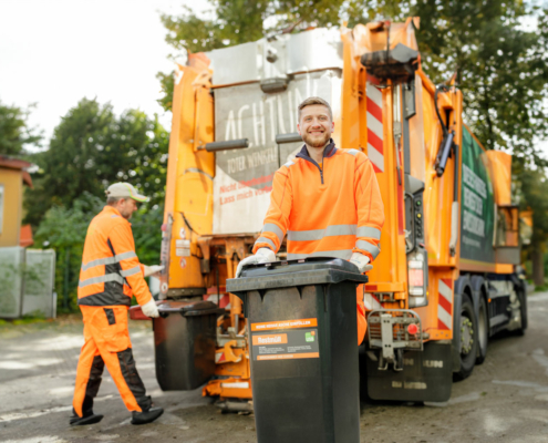 Zwei Männer holen den Müll ab. Sie stehen vor einem Müllwagen und sind orange gekleidet.