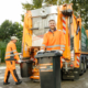 Zwei Männer holen den Müll ab. Sie stehen vor einem Müllwagen und sind orange gekleidet.