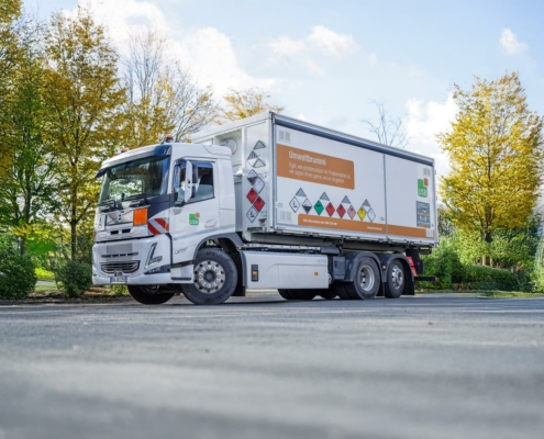 Der Umweltbrummi des USB steht auf eine aspahltierten Fläche, im Hintergrund sind Bäume. Der weiße Kastenwagen hat an der Seite mehrere bunte Warnschilder.