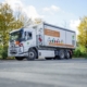 Der Umweltbrummi des USB steht auf eine aspahltierten Fläche, im Hintergrund sind Bäume. Der weiße Kastenwagen hat an der Seite mehrere bunte Warnschilder.