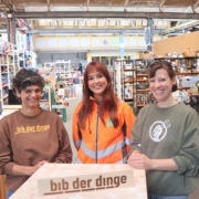 Drei Personen stehen in der bibliothek der dinge. Im Hintergrund sind die Regale zu sehen, in denen Verleihgegenstände liegen. Die Personen sind Dr. Najine Ameli Initiatorin der „bibliothek der dinge“, Kimberly Düpre von der USB Bochum GmbH und bib-Dozentin Bettina Strunk.