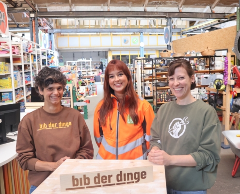 Drei Personen stehen in der bibliothek der dinge. Im Hintergrund sind die Regale zu sehen, in denen Verleihgegenstände liegen. Die Personen sind Dr. Najine Ameli Initiatorin der „bibliothek der dinge“, Kimberly Düpre von der USB Bochum GmbH und bib-Dozentin Bettina Strunk.