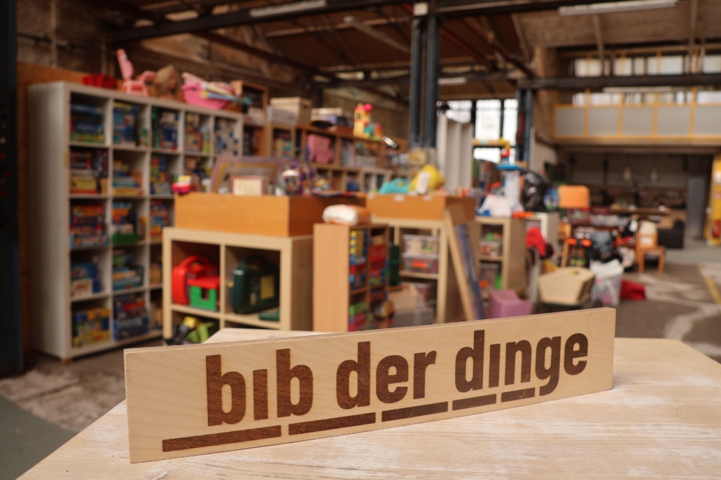Der Schriftzug der bib der dinge ist im Vordergrund zu sehen. Im Hintergrund sieht man Regal mit verschiedenen Verleihgegenstände.